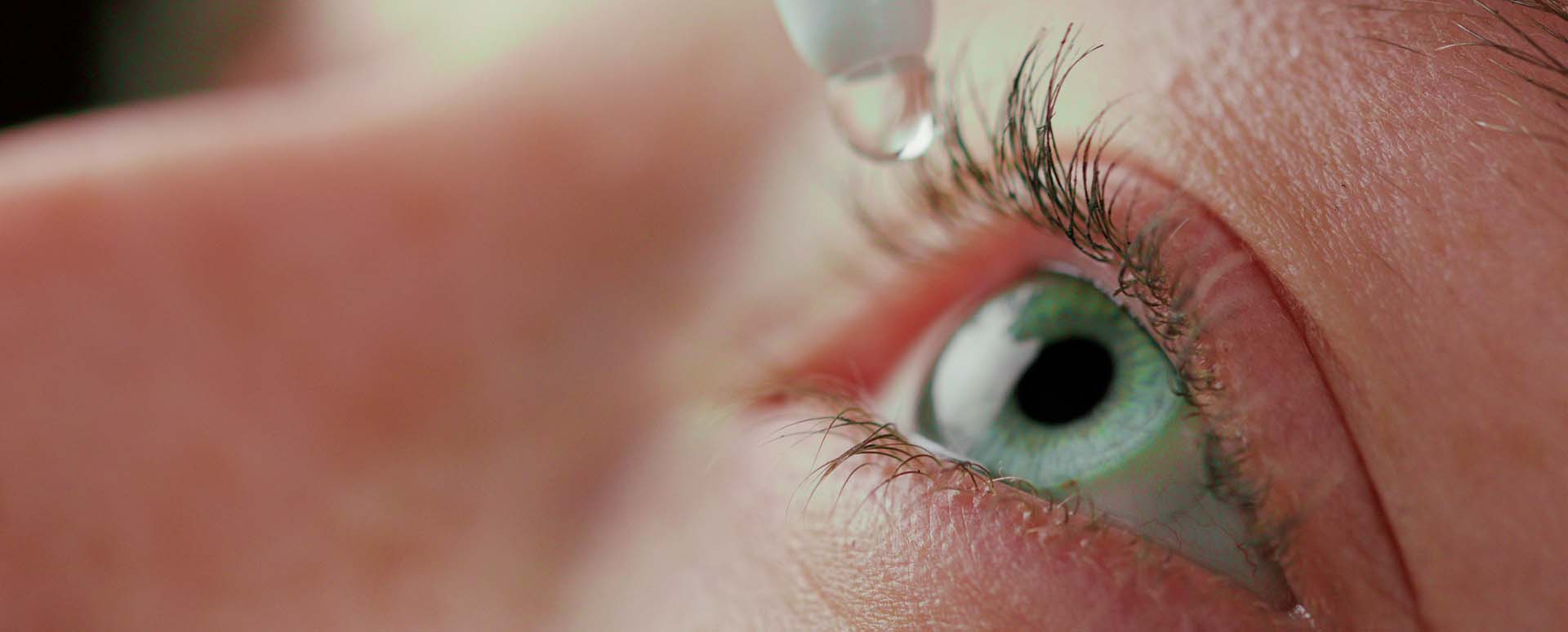 A tip of a white eye drop bottle with an eye drop above a agreen eye