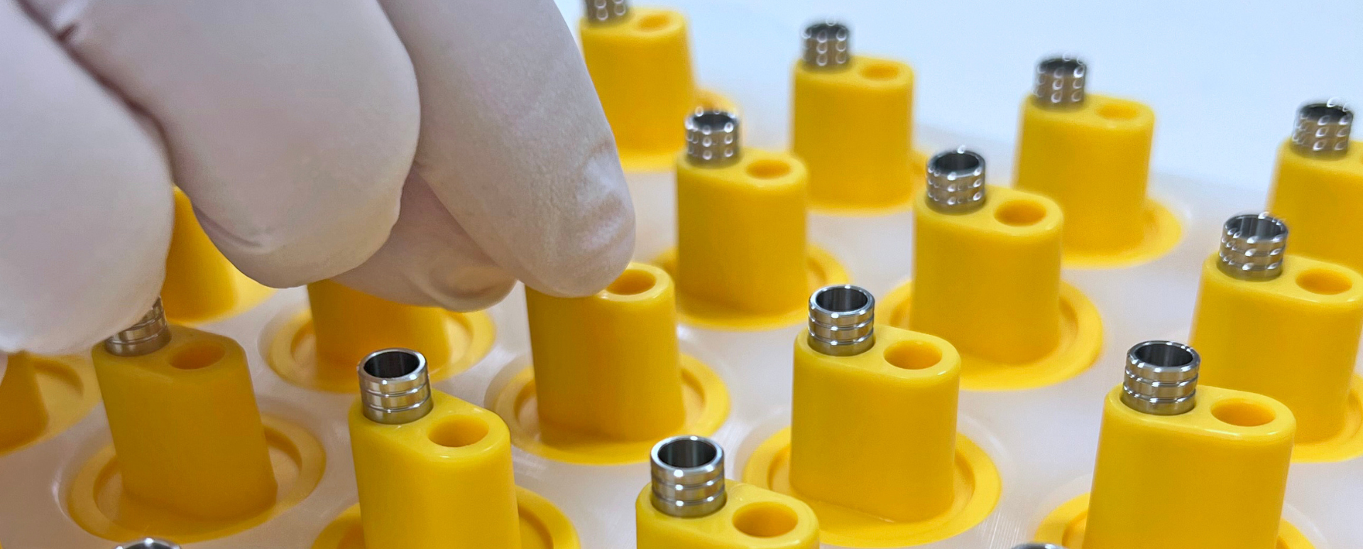 Shows a gloved hand assembling metal medical device components into a tray of yellow parts. 