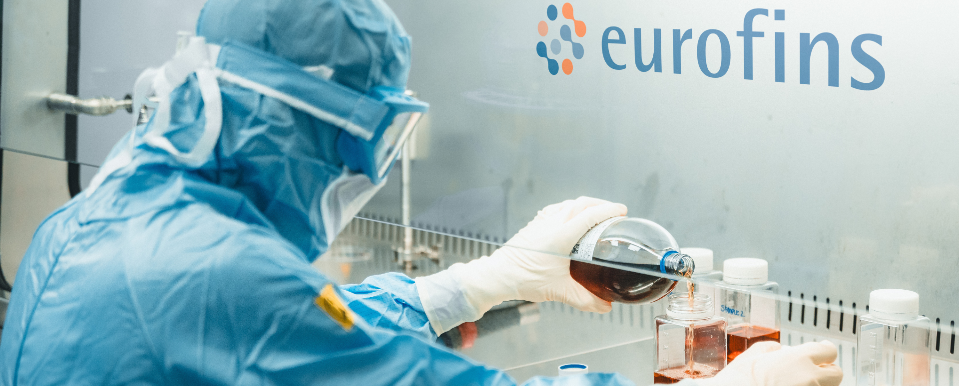 A laboratory worker clothed in a blue lab overall carrying out a test in a laboratory environment. Eurofins Logo on the wall.