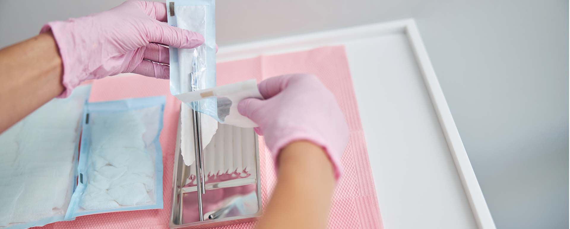 Two gloved hands are opening a plastic package containing a scalpel on a white table with a pink protective sheet