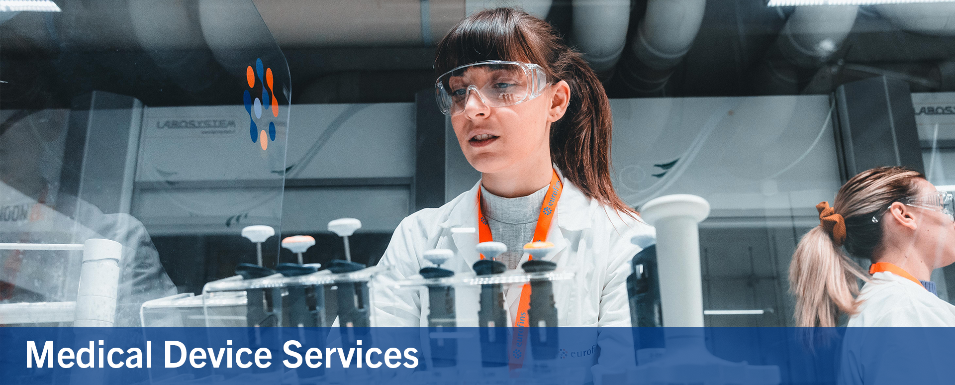 Two female scientists in a laboratory wearing safety goggles and lab coats, working with pipettes and medical devices. The image is related to medical device services, emphasizing precision and expertise in a clinical or research setting