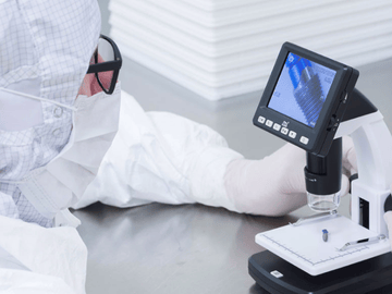 A laboratory worker in a clean room overall wearing a surgical mask and gloves, carrying out a test on a component of a medical device. 