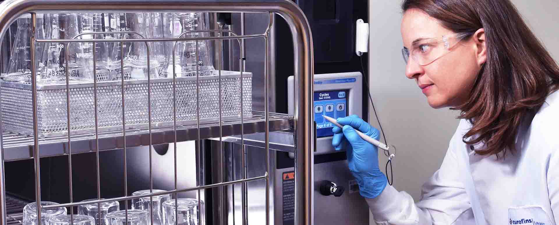A woman in a lab cost is holding a stylus in her gloved hand to adjust settings on an autoclabe being filled with two shelves of clear beakers in bins.