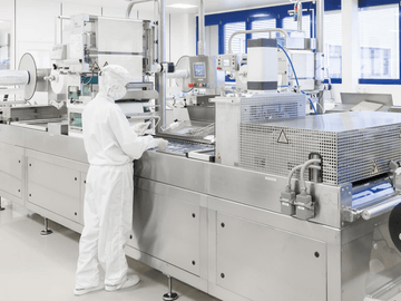 Shows a production worker in a clean room overall standing next to a blister production machine. 