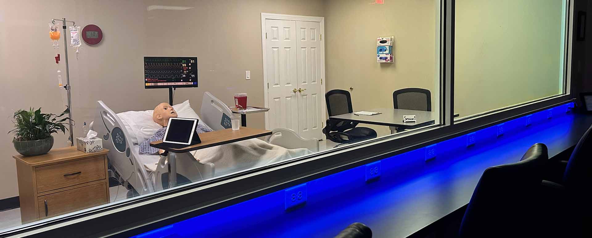 Chairs next to a window to view a lab simulation of a mannequin in a hospital bed with a night stand and IV pole