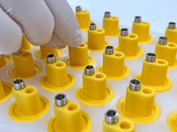 Shows a gloved hand assembling metal medical device components into a tray of yellow parts. 