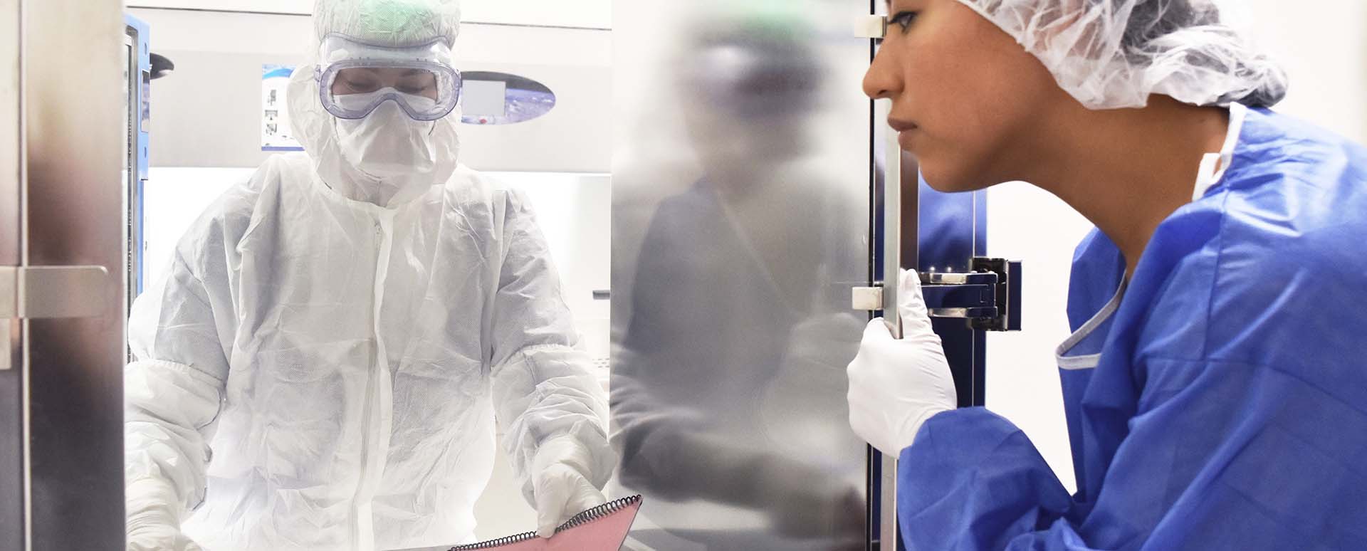 A person in white coverall with white gloves and laboratory safety goggles is handing a bound report with a red cover through a window with a door to a woman with white gloves, blue coverall and a hair net