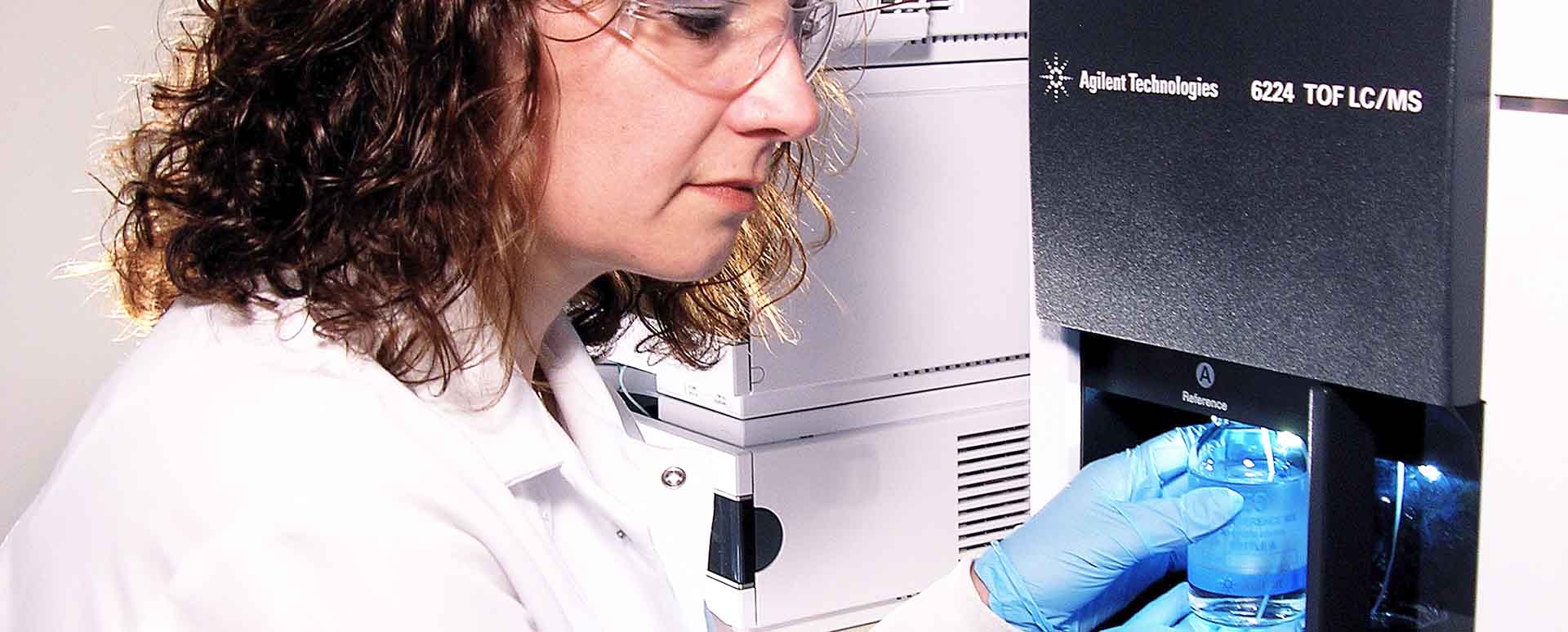 A brunette woman in a lab coat and glasses holds a sample at an Agilent 6224 TOF LC/MS with gloved hands.