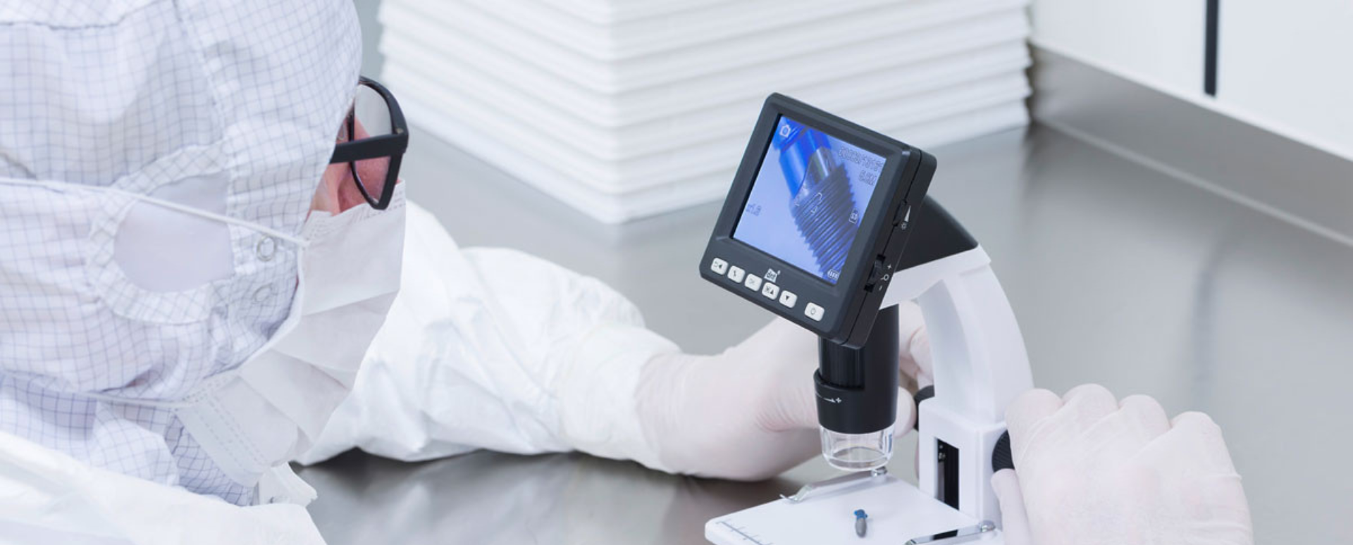 A laboratory worker in a clean room overall wearing a surgical mask and gloves, carrying out a test on a component of a medical device. 