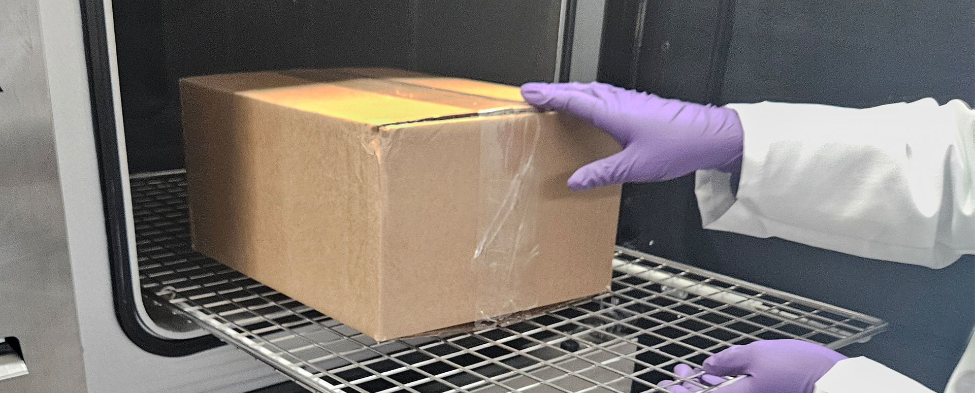 A Cardboard Box being placed in a steriliser by a person wearing a lab coat with one purple gloved hand on the box and one purple gloved hand on the chamber tray.