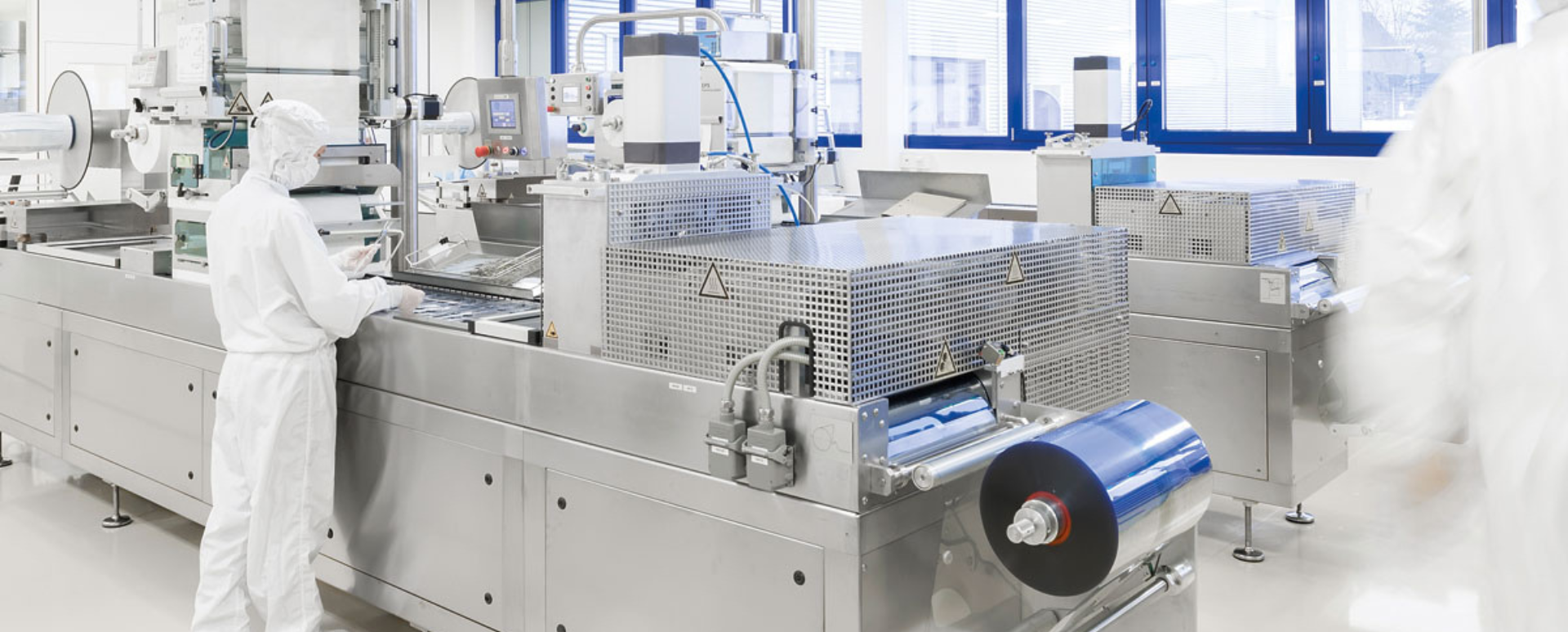 Shows a production worker in a clean room overall standing next to a blister production machine. 